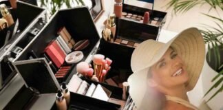 donna sorridente con grosso capello e una serie di trucchi alle sue spalle